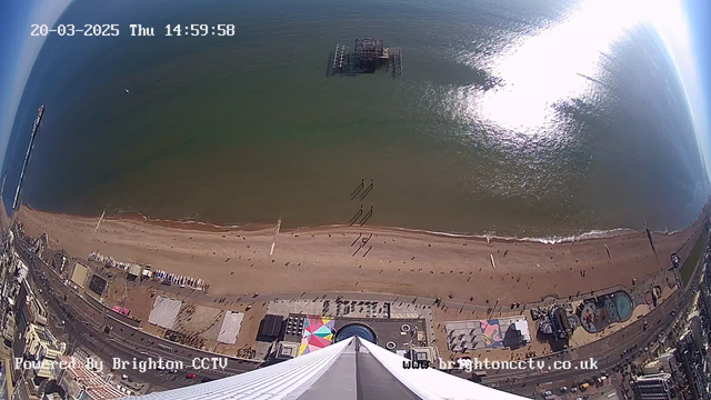 A bird's-eye view of a beach scene. The image shows a wide stretch of sandy beach with scattered people walking. To the left, there is a pier extending into the water. A large, abandoned structure is visible offshore. The water reflects sunlight, creating a bright area on the surface. The bottom of the image features a colorful beachside area with structures and attractions. The sky is clear and blue. A timestamp in the corner indicates the date and time.