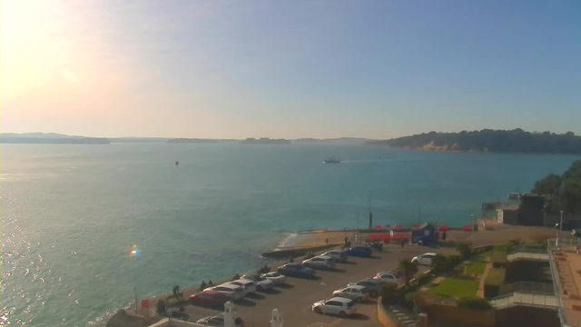 A sunny seaside view featuring calm, blue water and a distant shoreline with green hills. In the foreground, there is a parking lot filled with cars, while a small boat navigates in the water. Beyond the parking lot, a pathway leads to the water's edge, where some people are gathered near the shore. The sky is clear, and the sunlight glimmers on the water's surface.