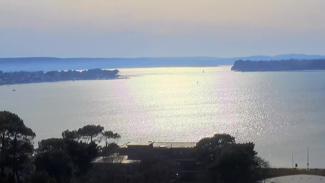 A serene waterfront view featuring a shimmering expanse of water reflecting hues of silver and blue, with distant hills and small islands visible on the horizon. In the foreground, trees outline the water’s edge, while a few boats can be seen floating in the calm water. The sky above is softly lit, indicating either early morning or late afternoon.
