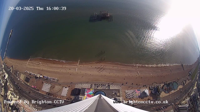 A wide aerial view of a beach and coastline, taken from a high vantage point. The image captures the sandy beach with numerous small figures of people walking along it. A wooden pier extends into the water, with some structures visible on the shoreline. The sea appears calm and reflects sunlight. There are colorful tents or structures near the beach area, and additional buildings and roads can be seen to the left. The image also includes a timestamp at the top showing the date and time of capture.
