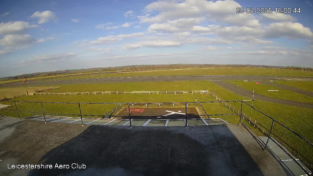A view from a webcam at the Leicestershire Aero Club shows a vast open landscape with a clear blue sky scattered with a few white clouds. In the foreground, a flat rooftop with a railing can be seen, while the background features a large grass field and a runway. Several white markers are visible on the ground, along with a red sign and white fencing lining the field.