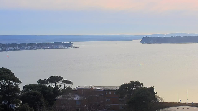 A view of a calm body of water stretching to the horizon, with patches of land and buildings visible along the shoreline. In the foreground, there are trees and the top of a building with a flat roof. The sky is a soft gradient of light colors, transitioning from blue to pale pink near the horizon. The water reflects these colors with a smooth surface, and a few boats can be seen in the distance.