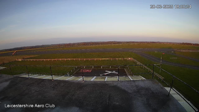 A view from a webcam at Leicestershire Aero Club shows an open field and runway area in the foreground. There are green grasses and a marked helipad with red and white stripes. In the distance, a flat landscape stretches toward the horizon under a clear sky, transitioning to soft hues of sunset. A white fence lines part of the area, and several taxiway markings are visible on the runway.