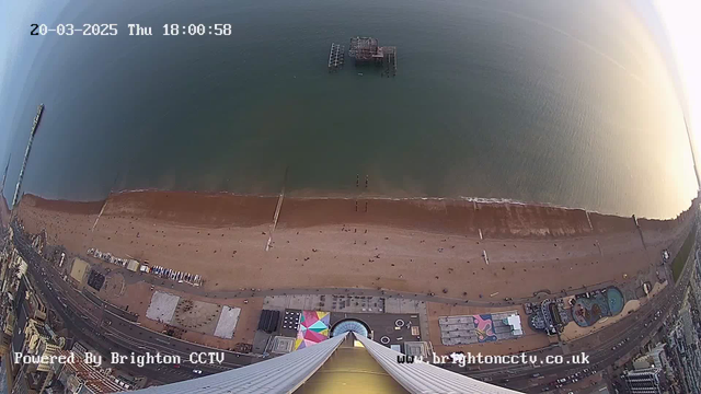 A high-angle view of a beach and ocean during the evening. The sandy shore is visible, with small dots of people scattered throughout. There is a pier extending into the water from the left side of the image, and a large, old wooden structure visible in the distance on the water. The beach area includes various buildings, some with colorful designs. A circular pattern is seen on the ground near the bottom of the image, suggesting an amusement park or similar attraction. The sky is illuminated with soft light, indicating sunset. The date and time are displayed in the top corner.