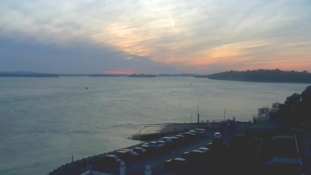 A serene view of a body of water during sunset. The sky displays soft, pastel colors of orange, pink, and blue, with wispy clouds. The water reflects these colors, creating a tranquil atmosphere. In the foreground, there are several parked cars along a road, with a small pier visible to the right. The landscape features distant hills on the horizon and a few small boats on the water.