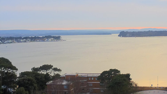 A panoramic view of a calm body of water under a soft, hazy sky. In the foreground, there are low buildings with dark roofs and some trees. The water reflects light, creating a gradient from gray to gold. In the distance, there are hills and a cove with boats moored along the shoreline. The overall atmosphere is tranquil and serene.