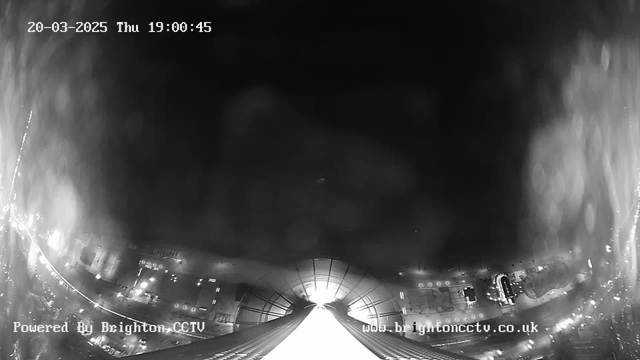 A black and white image captured by a webcam, showing a city skyline at night. The foreground features a downward view of a brightly lit path or road, leading away from the camera. In the background, there are faint outlines of buildings and streetlights that appear as small bright spots against the darker surroundings. The date and time at the top indicate March 20, 2025, at 19:00:45, while a watermark at the bottom left shows "Powered By Brighton CCTV."