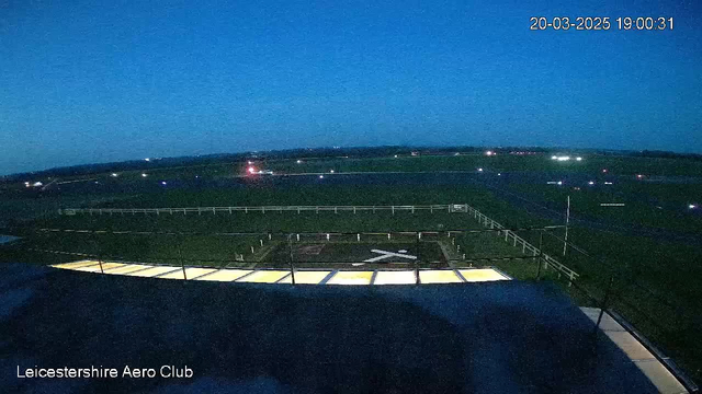 A twilight scene of an airport runway viewed from above. The foreground shows a section of the airport building with illuminated windows. In the middle ground, a grassy runway is bordered by a white fence, with a large white "X" marking a helipad. Distant lights flicker along the runway, indicating parked or preparing aircraft. The sky is a deep blue, suggesting evening time.