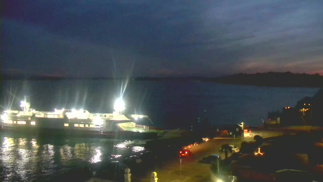 A dark scene shows a large ferry illuminated by bright lights, docked at a waterfront. Reflections of the lights shimmer on the water's surface. The sky is cloudy, transitioning from light to dark, indicating dusk. To the right, there is a winding road with parked cars and streetlights illuminating the area. The distant shoreline is faintly visible.