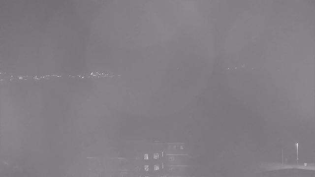 A hazy, monochromatic image shows faint lights in the distance, suggesting urban visibility through fog or mist. The lower part of the image features a dark structure, possibly a building, with some lit windows. The background is largely obscured, with only a hint of light breaking through the fog.