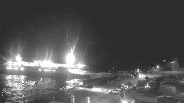 A nighttime scene featuring a large ferry docked at a port with bright lights illuminating the area. The water next to the ferry reflects the light. In the foreground, there is a parking area with several cars visible. The overall atmosphere is dark, with only the lights providing visibility. There are faint outlines of buildings and infrastructure in the background.