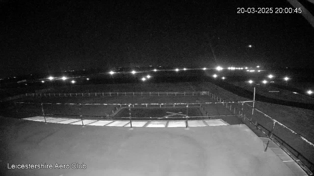 A dark scene with minimal lighting, showing the airfield of Leicestershire Aero Club at night. In the foreground, there is a flat surface, possibly a rooftop, with a low railing on the left side. The background features a large expanse, indicating an airfield with several distant lights scattered across the landscape, suggesting the presence of aircraft or vehicles. The time stamp at the top right indicates it is 8:00 PM on March 20, 2025.