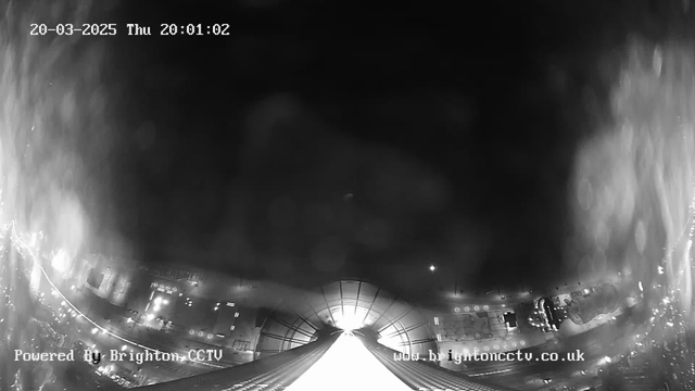 A black-and-white webcam image shows a view from a high point overlooking a cityscape at night. The scene is illuminated by various lights from buildings and streets below, creating a glowing pattern. In the center, a bright light source is visible, likely from a structure or street. The date and time in the top left corner indicate it is Thursday, March 20, 2025, at 20:01:02. The bottom left corner has text stating "Powered By Brighton CCTV." The overall atmosphere is nighttime with a slight foggy or misty effect.