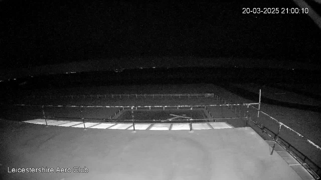 A dark scene from a webcam at Leicestershire Aero Club showing an empty airfield. The foreground features a flat surface with some illuminated patches, possibly from lights. In the background, faint lights are visible on the horizon, indicating a distant city or other structures. There is a low barrier along the side of the airfield, and the overall atmosphere suggests nighttime conditions. The timestamp is displayed at the top right corner, indicating the date and time.