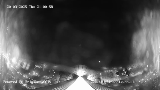 A black and white image from a webcam showing a view of a cityscape at night. The scene appears to be slightly blurred with reflections. In the foreground, a bright light source is visible, possibly indicating a road or pathway leading towards a building. The left side of the image includes illuminated structures, potentially vehicles parked along a street; some lights are also visible in the distance. The timestamp in the image indicates the date and time: March 20, 2025, at 21:00:58. A watermark at the bottom left indicates this is powered by Brighton CCTV.