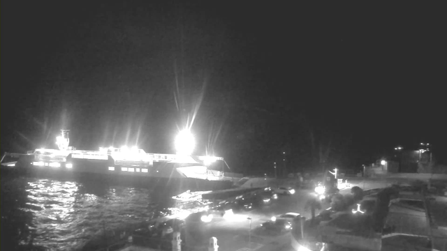 A night scene showing a large ferry docked at a harbor, illuminated by bright lights. The water is visible, reflecting the lights, and there are several parked cars along the waterfront. The background also features buildings with some lights on, creating a contrast against the dark sky.