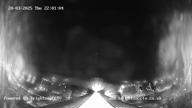 A black and white image from a high-angle webcam perspective showing a view of city streets and buildings at night. The scene is partially obscured by a haze or water droplets on the lens. The bottom of the frame displays a bright, illuminated structure, possibly a building or a bridge, leading into the distance. The date and time are shown in the upper left corner, indicating March 20, 2025, at 22:01:04, with a watermark for Brighton CCTV in the bottom left corner.