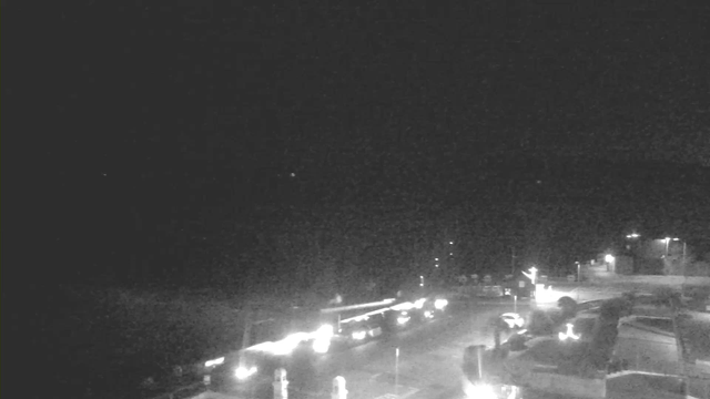 A dark scene captured from a webcam, featuring faint outlines of buildings and cars. The area is illuminated by scattered lights, with a few visible vehicles on a road. The background is largely obscured, suggesting proximity to water or an open space, while some light sources create a subtle glow in the night.