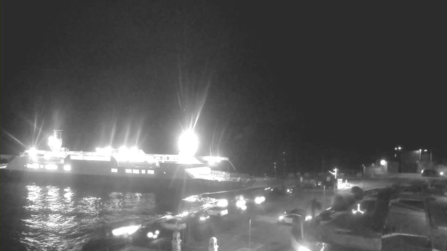 A dark night scene featuring a large ferry illuminated by bright lights, docked near a waterfront. The surface of the water reflects the lights from the ferry. Surrounding the area, there are several vehicles with headlights on, indicating movement. The shore contains various structures, including what appears to be a harbor or port area with infrastructure and trees nearby. The sky is mostly dark, emphasizing the bright lights of the ferry and vehicles.