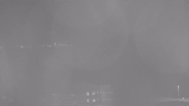A dimly lit scene shows a hazy view from a webcam. In the foreground, there are blurred outlines of a building with several windows. The background features a faint line of lights, suggesting a distant area, possibly a city skyline or shore. The overall atmosphere is foggy, limiting visibility and contrast.