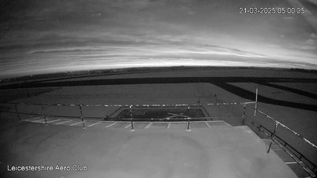 A view from a webcam on a cloudy early morning. The sky is dark with streaks of lighter gray, indicating dawn is approaching. A flat, open area is visible in the foreground, likely a runway or tarmac, with a white painted 'X' marking in the center. There is a railing in the foreground, and the distant horizon has a faint outline of what appears to be land or buildings. The overall image is monochrome and dimly lit.