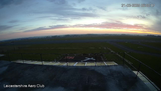 A wide-angle view from a rooftop overlooking an airfield at dawn. The sky is painted with soft hues of pink and blue as the sun begins to rise. In the foreground, a helipad marked with a white cross is visible, surrounded by green grass and a fence. The runway stretches out in the background, bordered by more grass and a slight fog that adds depth to the scene. The overall atmosphere is calm and serene, indicating early morning tranquility.