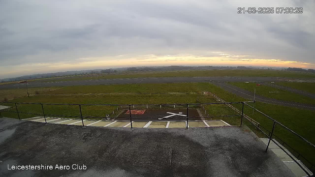 A view from a webcam overlooking a grassy airfield with a runway. The sky is overcast with hints of orange indicating sunrise. In the foreground, a balcony railing is visible, along with a white cross marking a landing area. There is a red marking visible on the ground, and in the distance, low hills are obscured by mist. The scene conveys a tranquil morning atmosphere at the Leicestershire Aero Club.