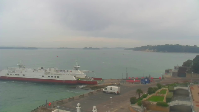 A cloudy sky looms over a body of water with several islands visible in the distance. A white and red ferry is docked at a harbor, with a gray van parked nearby. In the foreground, there are landscaped areas with small shrubs and pathways, while the background features more greenery and a steep coastline. Red obstacles are arranged along the dock area. The overall atmosphere suggests a calm but overcast day.