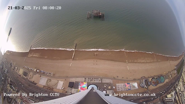 A wide aerial view of Brighton beach during the morning. The scene includes the coastline with a sandy beach bordered by the sea. Several piers extend into the water, with one partially submerged pier visible in the middle distance. In the lower part of the image, there are structures, including colorful beach huts and amusement rides, along the beachfront. The sky is mostly clear with a slight reflection of sunlight on the water. The date and time are displayed at the top of the image.