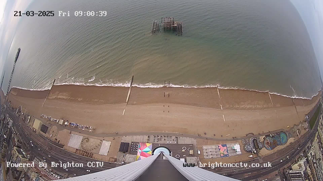 A view from a high vantage point looking down at a sandy beach with a gentle wave lapping at the shoreline. Several people are walking on the beach. The ocean is calm with an old, partially submerged pier in the distance. The area includes colorful beachside attractions and structures, with geometric patterns visible on some surfaces. A cloudy sky is above, and the scene is framed by a tall building in the foreground.