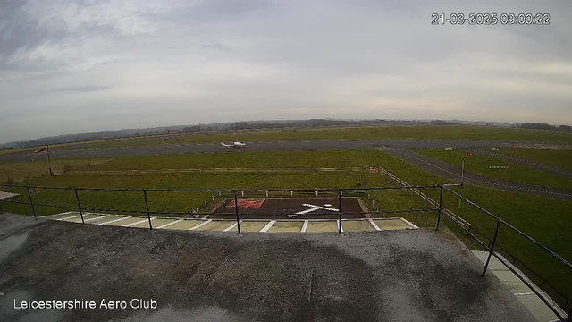 A view of a grassy airfield from a raised platform at the Leicestershire Aero Club. In the foreground, there is a railing with a concrete surface beneath. To the right, a small aircraft is taxiing on the runway, which is marked with white lines. There are scattered patches of grass and an 'X' marking on the ground, indicating a helipad. The sky is overcast with gray clouds, and the landscape in the background features distant hills.