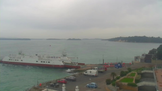 A chain ferry is docked at a shoreline, with its bow facing the camera. The water is calm and has a bluish-green hue. In the distance, various small islands are visible under a cloudy sky. On the land, there is a parking lot with several vehicles, including a white van and a red car. The foreground features landscaped areas with a few plants and trees. A dock with additional structures is seen to the right of the ferry.