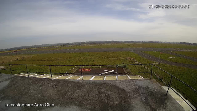 A view from a webcam showing the Leicestershire Aero Club. In the foreground, there is a rooftop railing, and below, a grassy area with a large "X" marked in white on the ground. The background features a runway, partially paved, leading into an expansive green field. The sky is mostly cloudy, giving a soft diffused light. The date and time stamp at the top indicates March 21, 2025, at 09:59:56.
