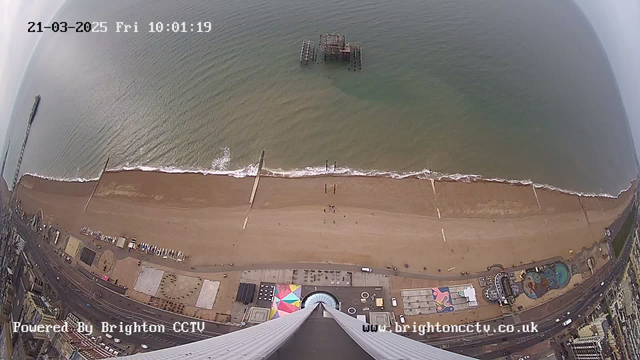 Aerial view of a sandy beach with gentle waves lapping at the shore. In the distance, there is a partially submerged pier structure. The beach is mostly empty, with a few people walking along the shore. Below, various colorful patterns on the ground can be seen, possibly from a fairground or amusement area. The ocean is a mix of greenish-blue hues, and the sky above appears overcast. Time and date are displayed in the top left corner.