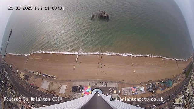 A high-angle view of a sandy beach along a coastline, with waves lapping at the shore. In the distance, remnants of a pier emerge from the water. Various people are scattered along the beach, and buildings can be seen at the bottom of the image, including colorful structures and amusement attractions. The sky above is partly cloudy. The time stamp on the image shows it is Friday, March 21, 2025, at 11:00:31.