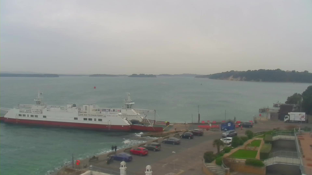 A large ferry is docked at a harbor, with a mix of cars parked nearby. The water is calm but slightly choppy, and the sky is overcast with gray clouds. In the background, there are hills with trees and a small shoreline. There are people visible near the ferry and around the parking area. Bright red and blue structures are seen along the dock.