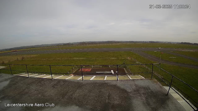 A wide view of a grassy airfield under a cloudy sky. The foreground shows a flat rooftop with a railing, and in the distance, there are marked runways. On the right side, a small aircraft is preparing for takeoff. The landscape is open, with a few trees and a fence bordering the airfield.