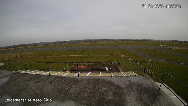 A view from a high vantage point overlooking an airfield. The foreground features a railing with a concrete surface. Below, there is a grassy area with a small section of red and white markings, possibly indicating a takeoff area. The runway extends into the distance with several landing lights visible along its sides. The sky is overcast with a pale gray tone, suggesting a cloudy day. In the far background, there are distant hills or landforms. The overall scene conveys a sense of an active aviation space.