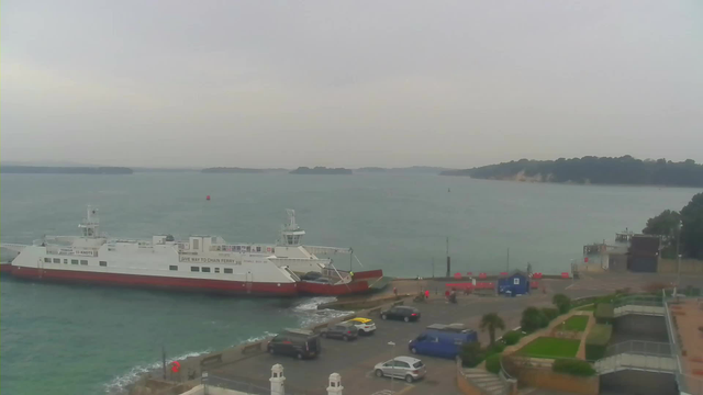 A ferry is docked at a terminal by the water, with waves gently lapping at the shore. Several vehicles are parked near the terminal, and a few people can be seen in the vicinity. In the background, there are islands and a cloudy sky, creating a muted atmosphere. The overall scene conveys a coastal setting.