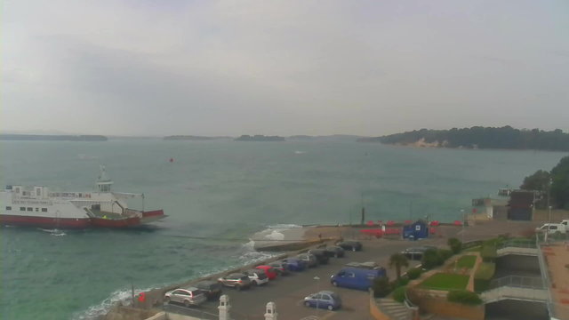 A coastal scene featuring a ferry approaching a dock. The water is choppy and appears blue-green under a cloudy sky. In the foreground, there is a parking area with several cars in various colors, including red, blue, and black. To the right, there are some buildings and greenery, along with a blue structure near the waterfront. Small boats can be seen in the distance against a backdrop of islands.