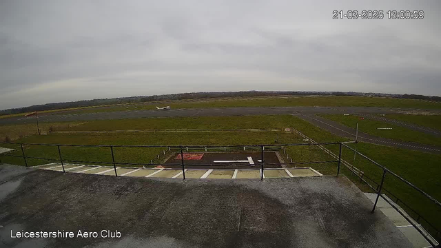 A view from a webcam situated at the Leicestershire Aero Club. The foreground features a flat surface with a gray texture, including white lines and a rectangular area with a red patch. In the middle distance, a runway stretches across the image, bordered by green grass on either side. A small aircraft is seen taking off or landing on the runway. The sky above is gray and overcast. The timestamp in the top right corner indicates the date and time of the image.