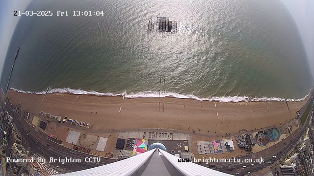 Aerial view of a beach on a sunny day. The scene shows the sandy shore lined with a few people and beach items. Waves gently crash onto the beach, where several wooden piers extend into the water. In the distance, an abandoned structure is partially submerged in the ocean. The foreground includes colorful patterns on a pavement, alongside a large circular structure. The water appears calm with a reflective surface, and the sky is clear. The image is timestamped and labeled as powered by Brighton CCTV.