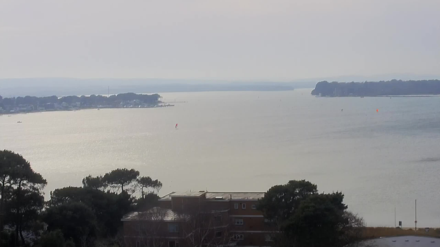 A calm body of water stretches into the distance, reflecting soft light with a silvery hue. In the foreground, there are dark green trees on the left, and a low building partially visible beneath them. Across the water, small boats are scattered, with a few noticeable red and orange markers. The horizon features a gentle, undulating shoreline with hazy outlines of land. The sky appears overcast, creating a muted atmosphere.