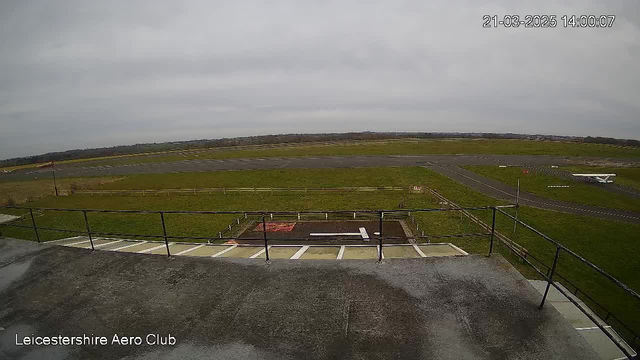 A view from a webcam shows an expansive grassy area with a runway. The sky is overcast with gray clouds. In the foreground, there is a railing along a flat surface, indicating a rooftop or balcony. In the middle ground, a small section of the runway is visible with a white plane parked to the right. Several wooden fences enclose parts of the grass area. The image is timestamped, indicating the date and time of capture.