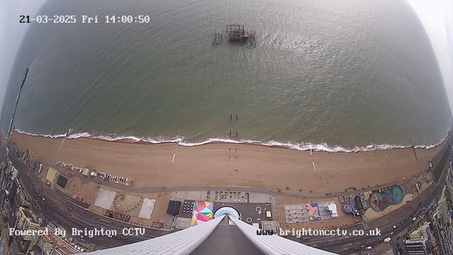 Aerial view of a beach and ocean taken from a high vantage point. The shoreline features gentle waves lapping against sandy beach, which is lined with people. Below, there are buildings, some with colorful patterns, scattered along the beach, including an amusement area with a pool visible. An old pier extends into the water at a distance. The sky appears cloudy but bright. The date and time are displayed in the top left corner.