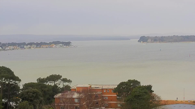 A view of a tranquil body of water surrounded by land. In the foreground, there are trees and a reddish-brown building with multiple windows. In the middle distance, a shoreline with various structures, likely homes or businesses, lines the water's edge. There are small boats visible on the water, and in the background, low hills fade into the misty sky, which is overcast. The overall scene conveys a calm, subdued atmosphere.