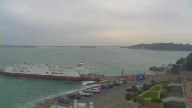 A view of a harbor on a cloudy day. In the foreground, there is a large white and red ferry docked at a pier. Several vehicles are parked along a road next to the water. The sea is choppy, with gentle waves. In the background, there are small boats in the water and islands visible in the distance. The sky is overcast, creating a muted light over the scene.