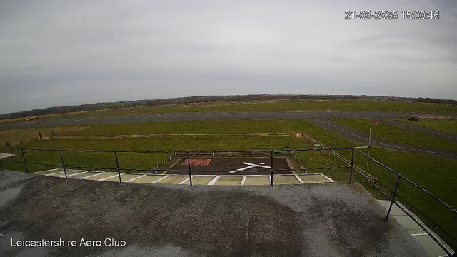 A view from a webcam showing a large open area likely representing an airfield. The foreground features a balcony or ledge with a railing. Below, there is clearly marked runway space with a large 'X' symbol on the ground. In the distance, patches of grass and some low fencing are visible along the perimeter of the airfield. The sky overhead is overcast, with a smooth gray appearance. The image is timestamped, indicating it was taken in the afternoon.