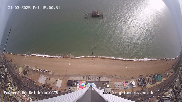 A high-angle view of a beach and the ocean, taken from a webcam. The sandy shore is visible with several people walking near the water's edge. To the left, a pier extends into the ocean, while an old structure is seen in the water. The beach features a colorful parasol and some beach equipment. The shoreline curves gently, and the ocean appears calm with gentle waves. The image includes a timestamp and a watermark indicating the source.