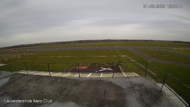 A view from a webcam located at an aeroclub shows a large, open grass area that is part of an airport runway. In the foreground, there is a flat rooftop with a railing. Below, a white "X" shape is marked on the asphalt, possibly indicating a landing spot. The sky is overcast with gray clouds, and the horizon features a distant line of trees and fields. A small aircraft is parked on the tarmac, while other runway markings and signs are partially visible in the background.